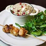 Close up of griddled chicken skewers on a plate, with a bowl full of couscous salad with feta and pomegranate in the background