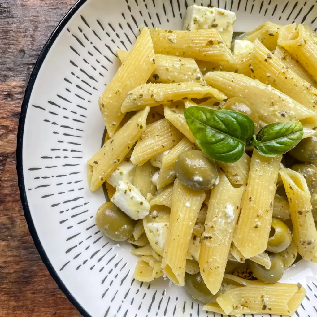 Cooked pesto pasta with olives and feta in a white bowl, topped with basil