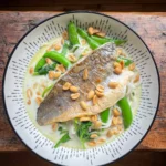 fried sea bass on top of a bowl of noodle soup topped with peanuts