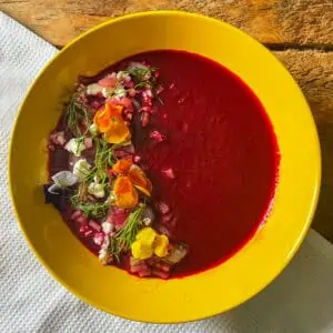 Bright red cold beetroot soup in a yellow bowl topped with feta, dill, onions and edible flowers